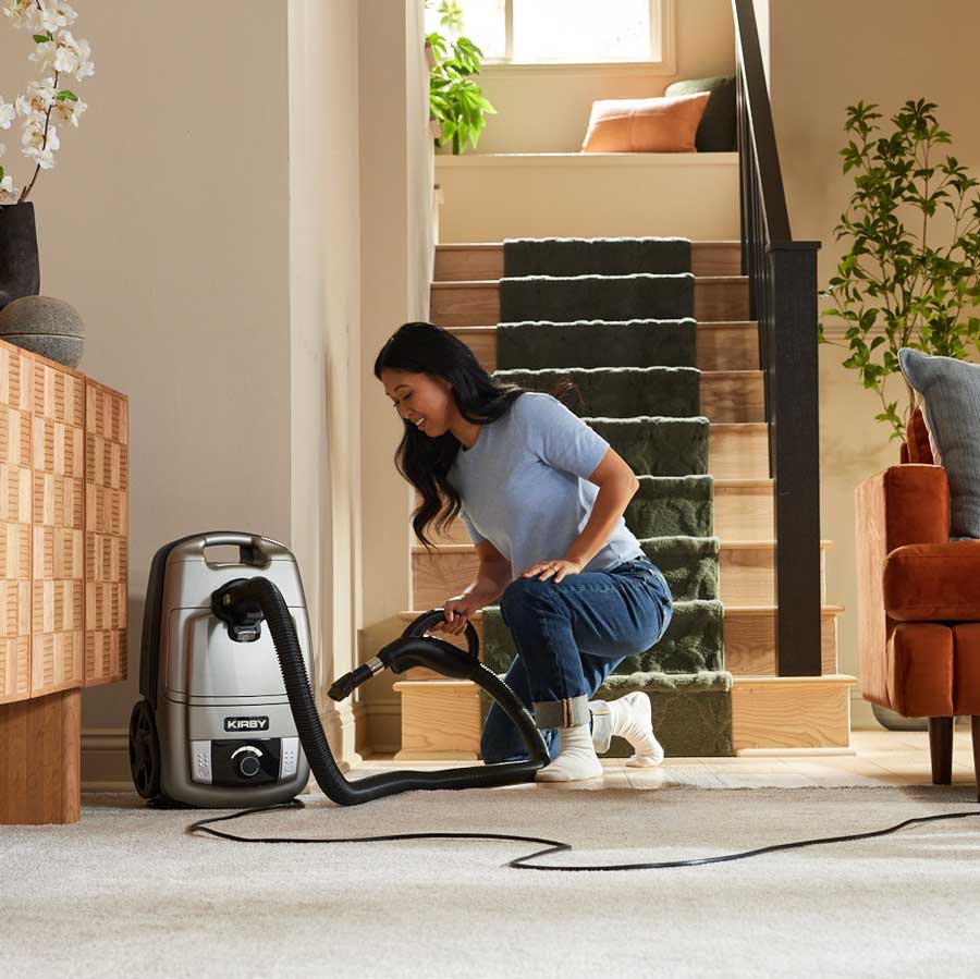 A woman in a home interior kneels beside a platinum Kirby Signature Series C530 canister vacuum, using a hose to clean a light-colored carpet.