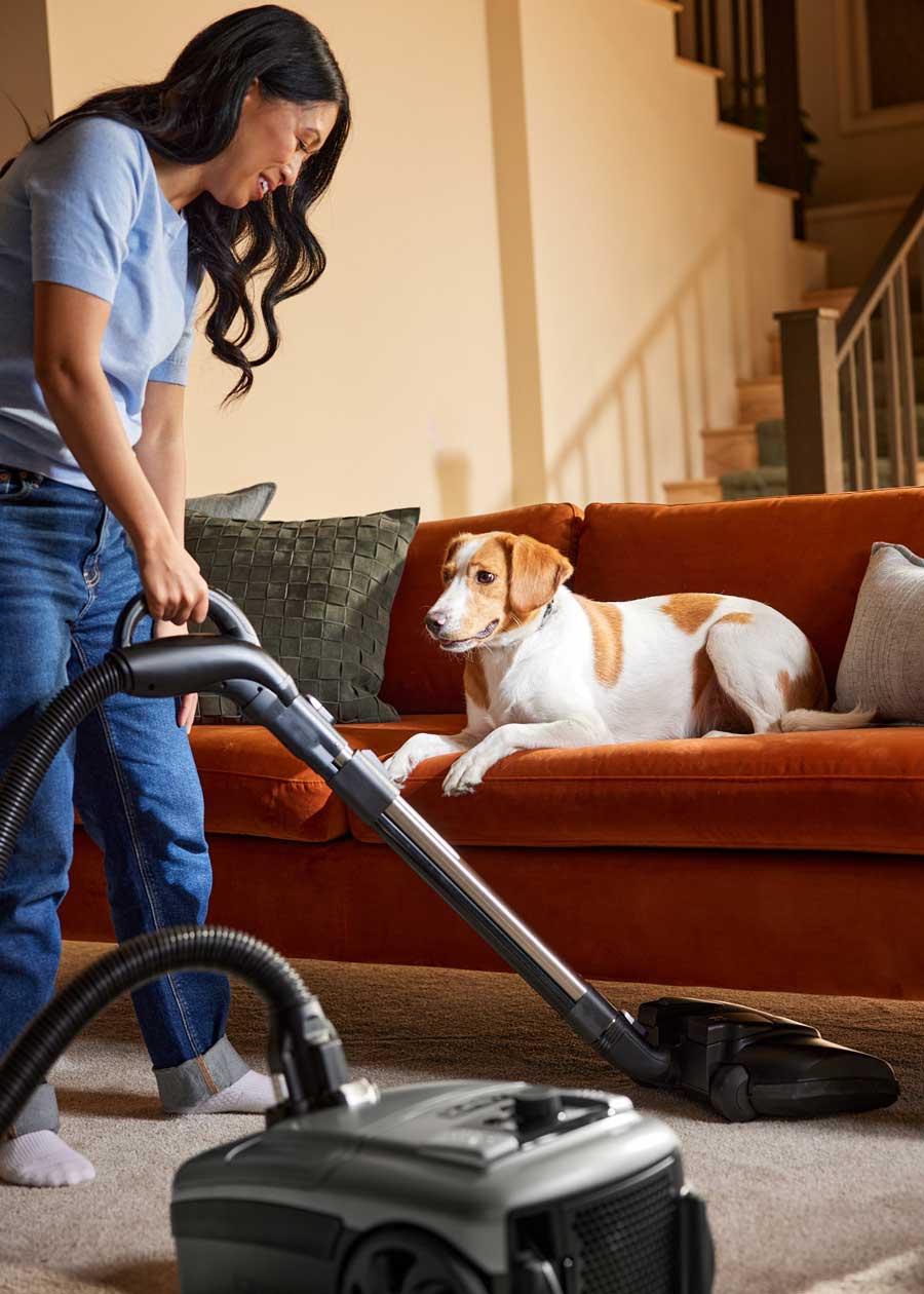 A woman uses a Kirby Signature Series C550 canister vacuum on a carpeted floor in a living room, with an orange and white dog lying on the couch.