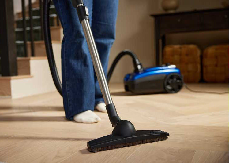 A person wearing blue jeans and white socks uses a Kirby Signature Series V530 canister vacuum cleaner with a black wand and floor head to clean a light wood herringbone floor in a home interior setting.