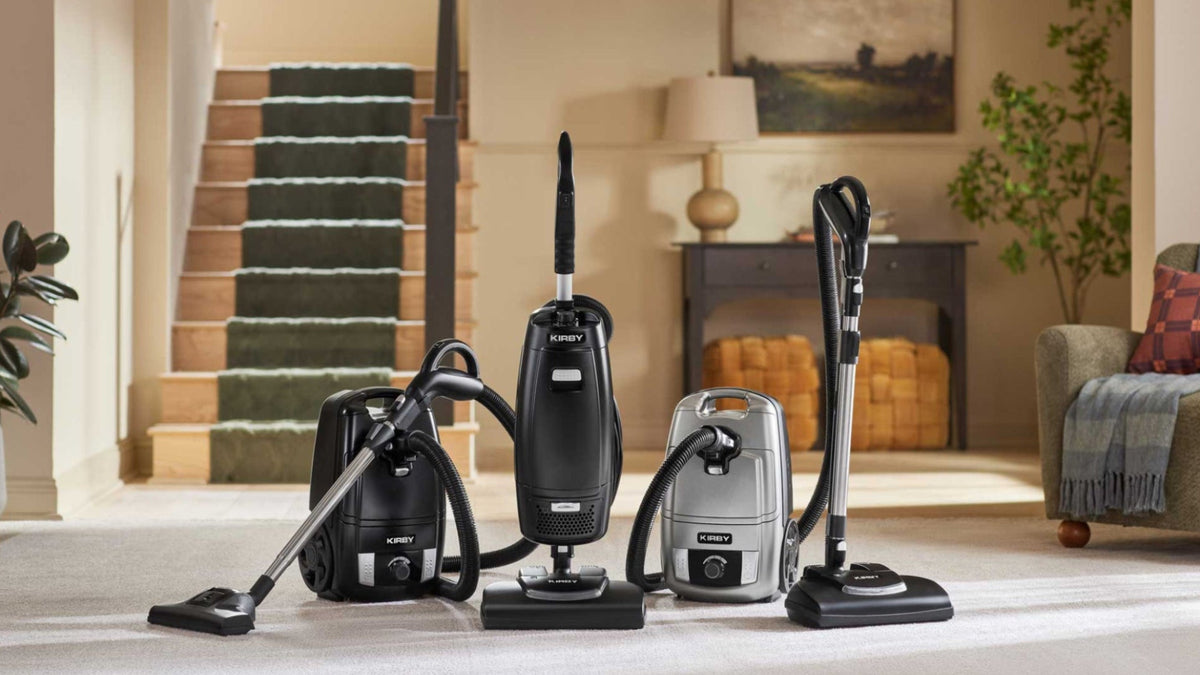 A lineup of three Kirby Signature Series vacuum cleaners in black and silver sit atop a light-colored carpet in a beautifully styled living room with a staircase in the background.