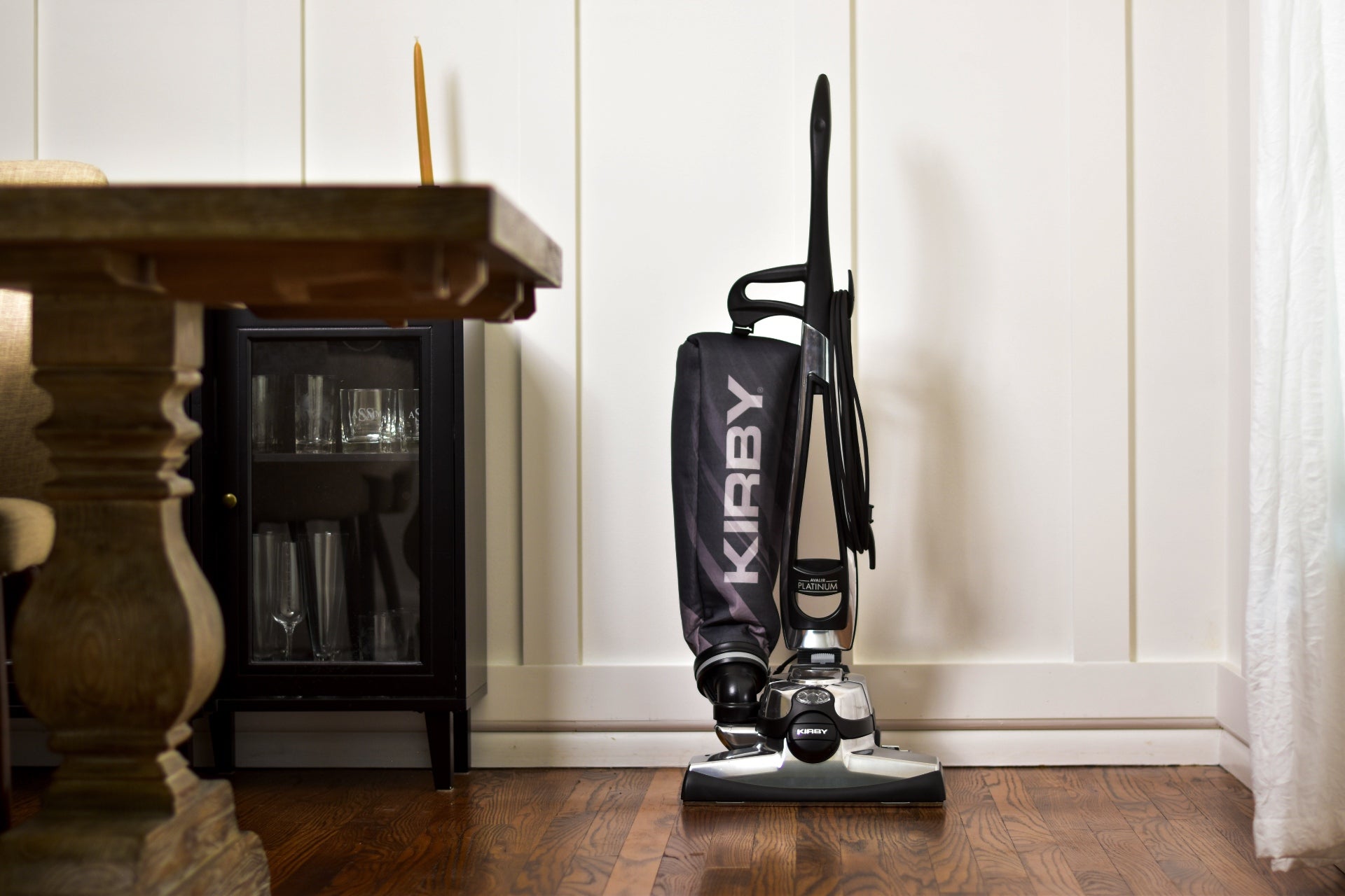 Black Kirby vacuum cleaner against a white paneled wall with a dark wooden cabinet and decorative items.