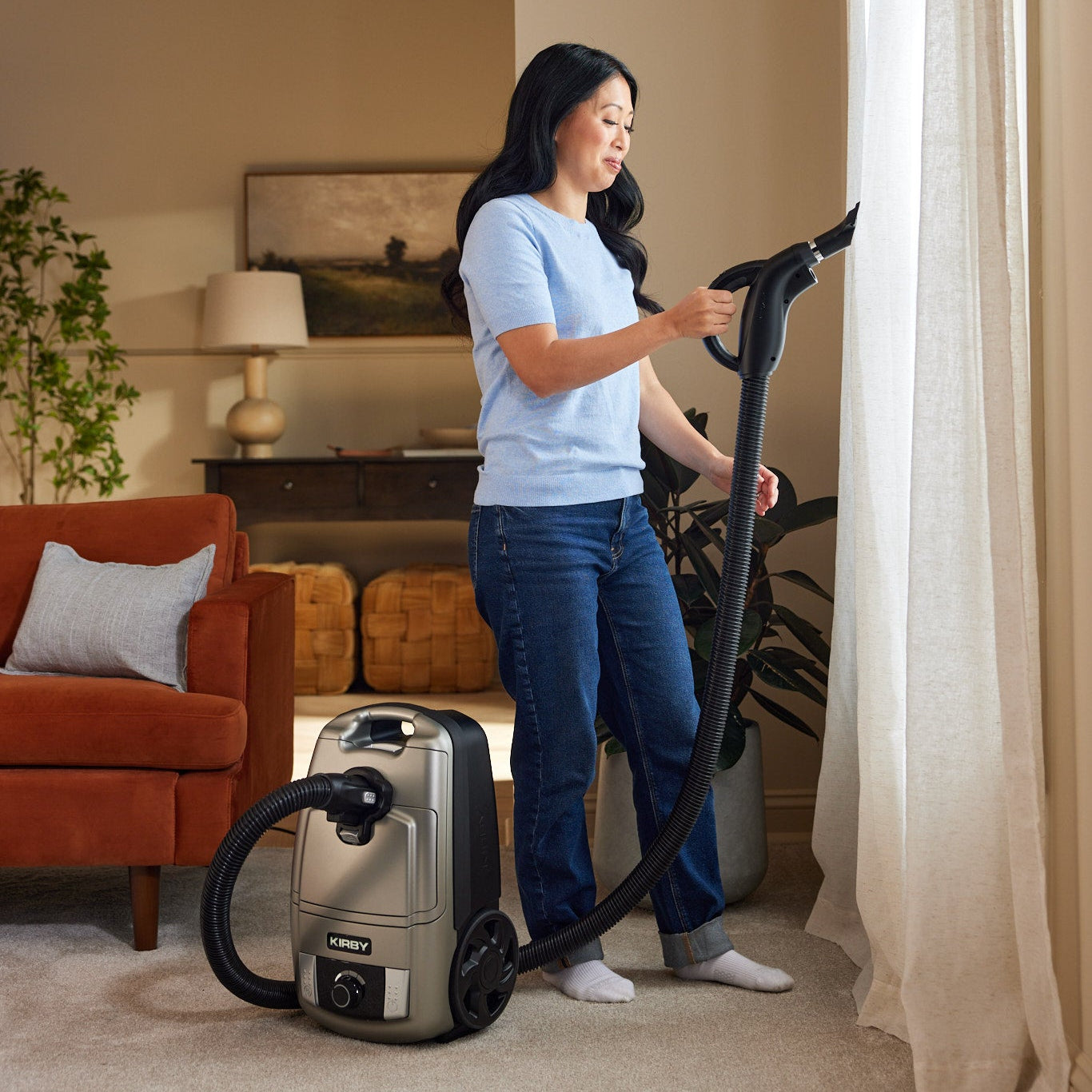 A woman in a living room uses a platinum Kirby Signature Series C550 canister vacuum cleaner with a hose, positioned next to a window with sheer curtains.