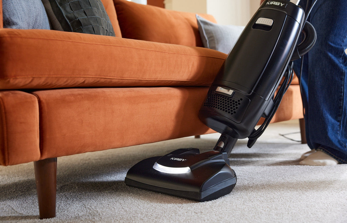 A black Kirby Signature Series V530 upright vacuum with a multi-surface Power Nozzle, shown being used under an orange couch on a light carpet.