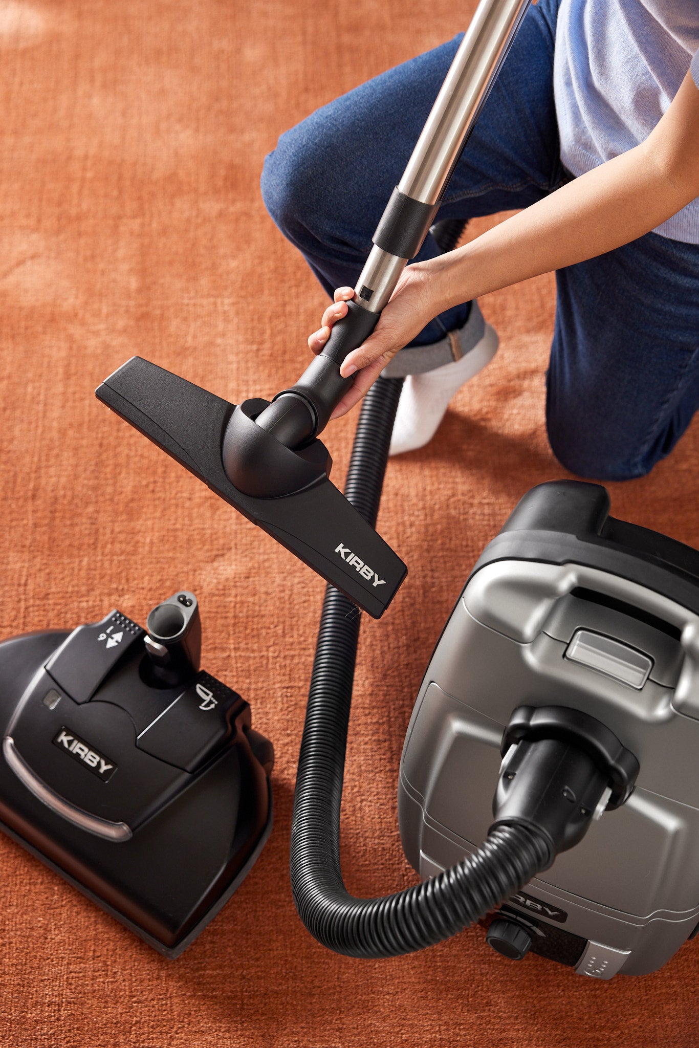 The Power Nozzle of a Kirby Signature Series C550 canister vacuum cleaner is removed and replaced with a floor nozzle, attached to a rod and flexible hose. Woman kneeling on orange carpet.