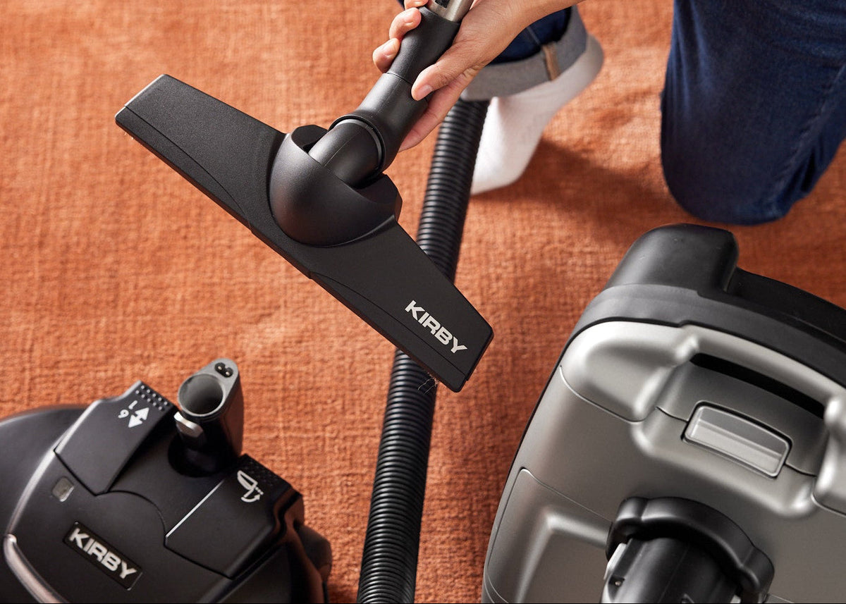 The Power Nozzle of a Kirby Signature Series C550 canister vacuum cleaner is removed and replaced with a floor nozzle, attached to a rod and flexible hose. Woman kneeling on orange carpet.
