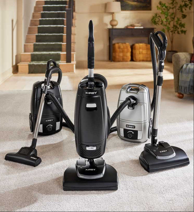 Three Kirby Signature Series vacuums in black and silver are displayed on a light beige carpet in a modern living room setting with a staircase in the background.
