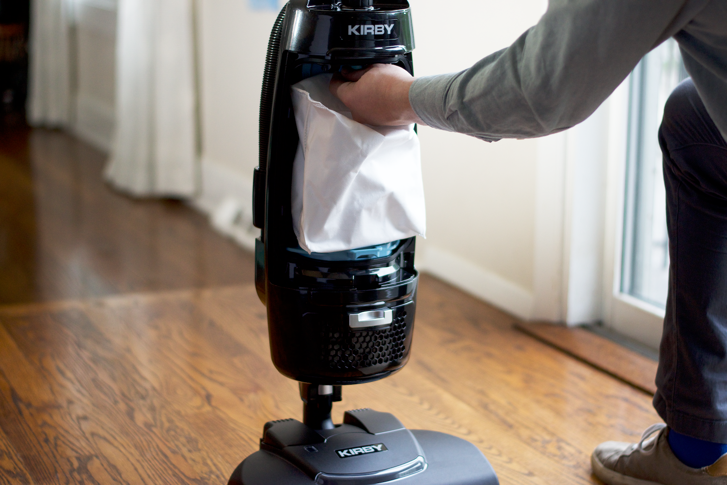 A person changing a PureAir Dual-HEPA filter bag within a Kirby Signature Series V530 upright vacuum cleaner atop a hardwood floor.