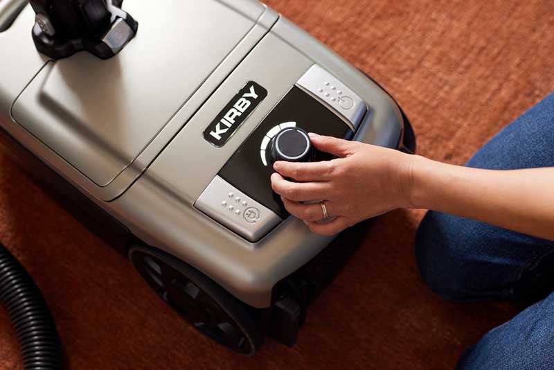 A close-up of a Kirby Signature Series C550 canister vacuum being operated on a brown carpet by a person whose hand and leg are visible.