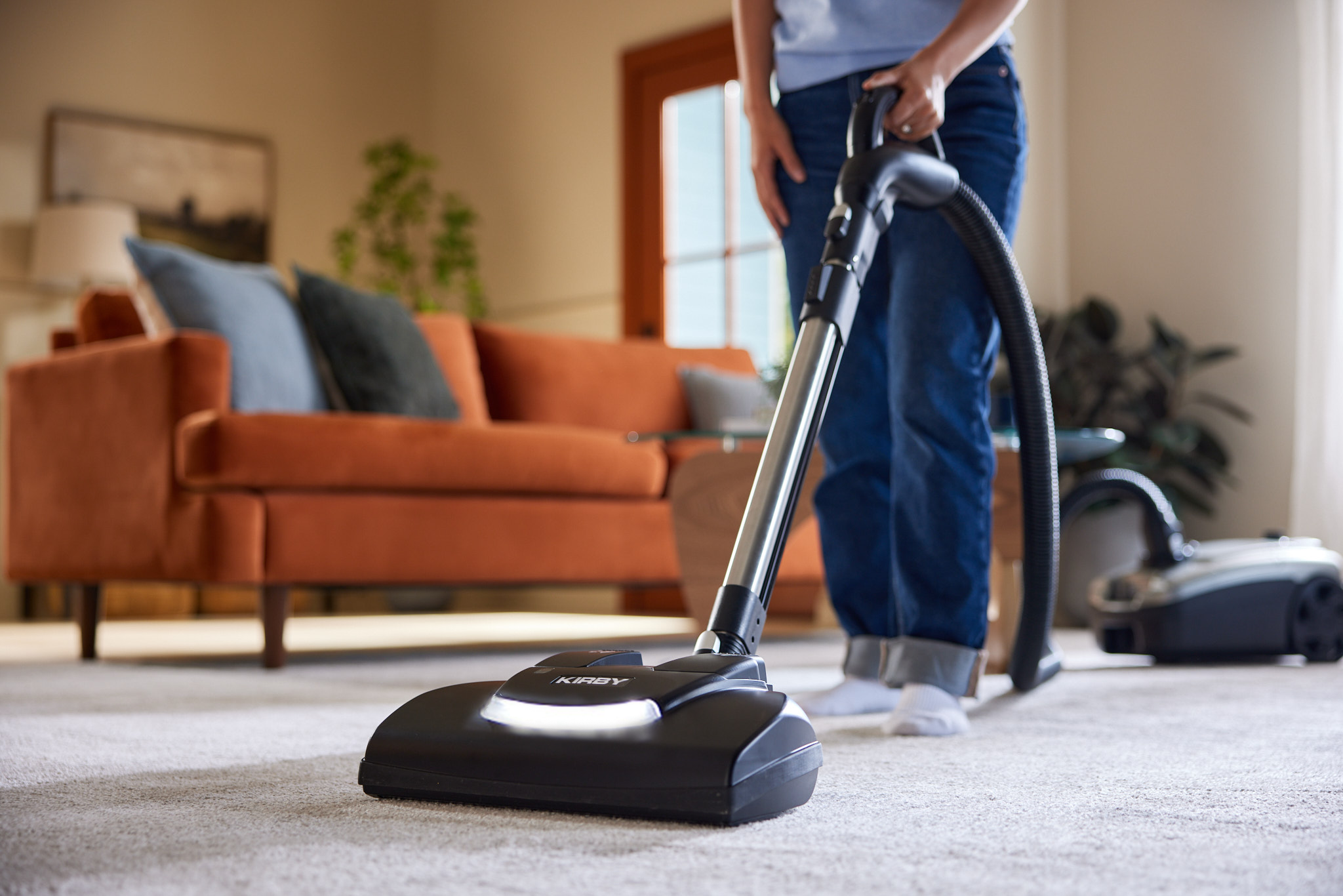 A woman using a black and silver Kirby Signature Series C550 canister vacuum on a light-colored carpet in a living room with an orange sofa in the background.