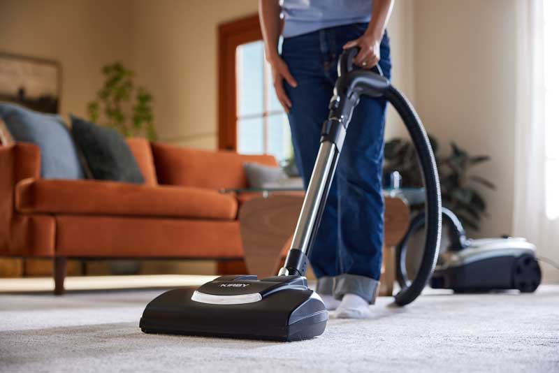A woman uses a Kirby Signature Series C550 canister vacuum cleaner on a light beige carpet in a modern living room with an orange sofa in the background.