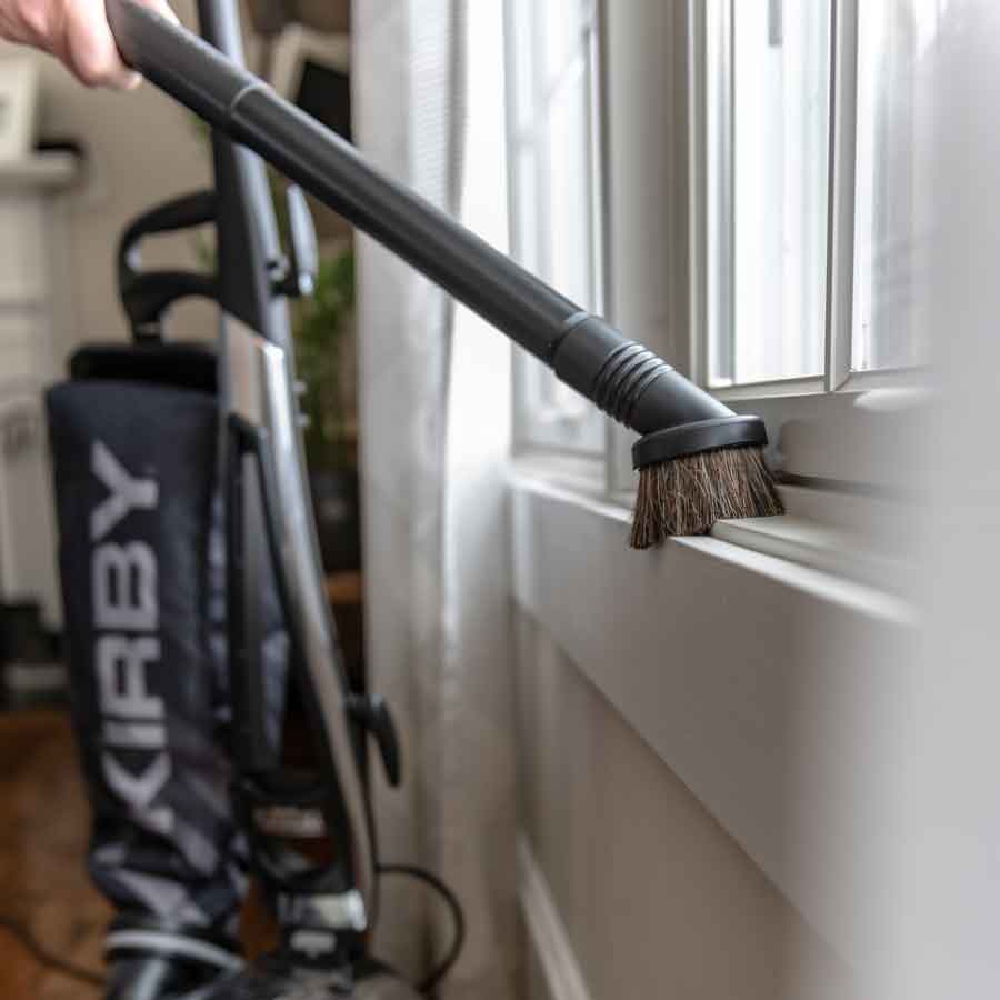Kirby vacuum with a black and silver design, using a dusting brush attachment on a window sill.