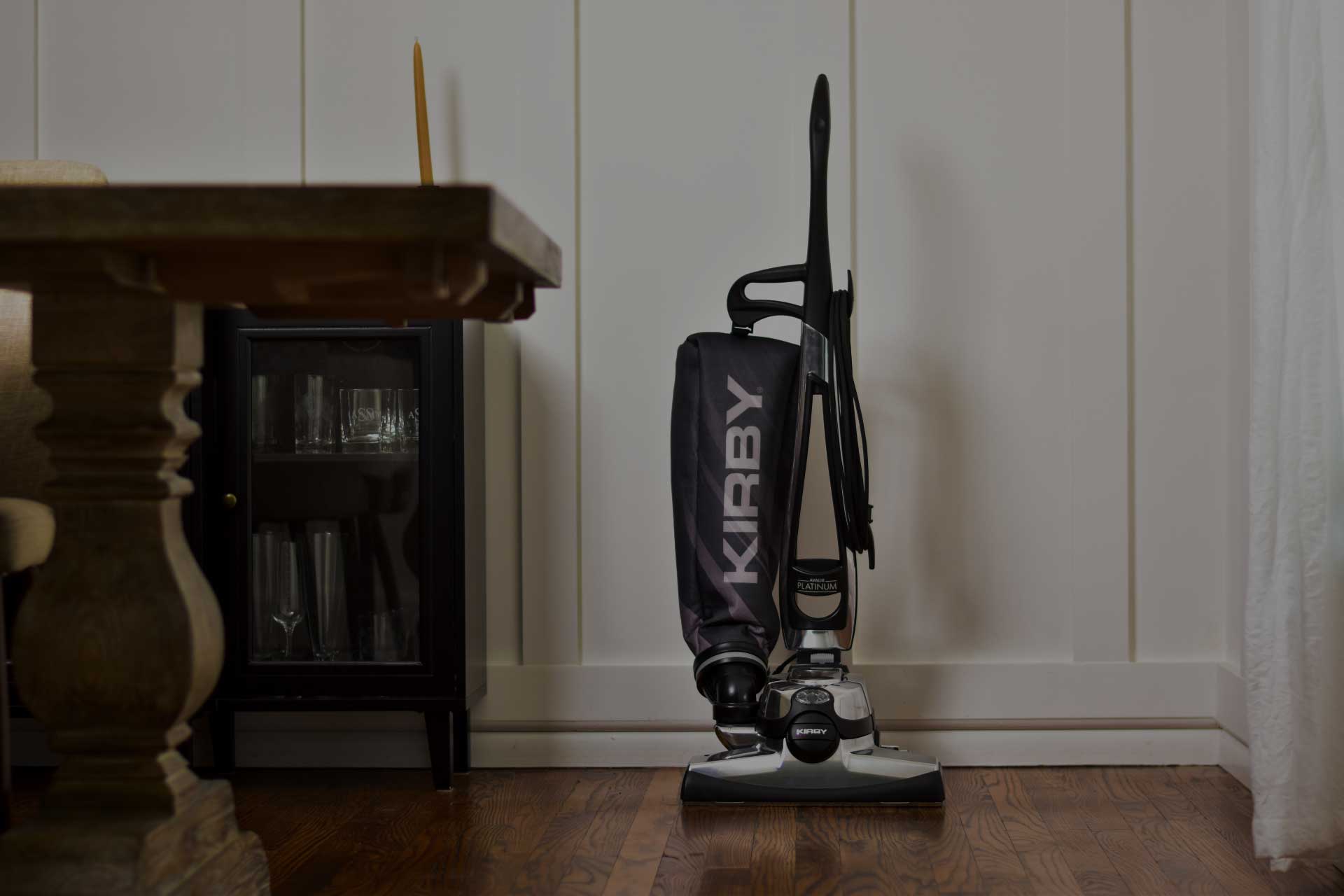 Black Kirby vacuum cleaner against a white paneled wall with a dark wooden cabinet and decorative items.