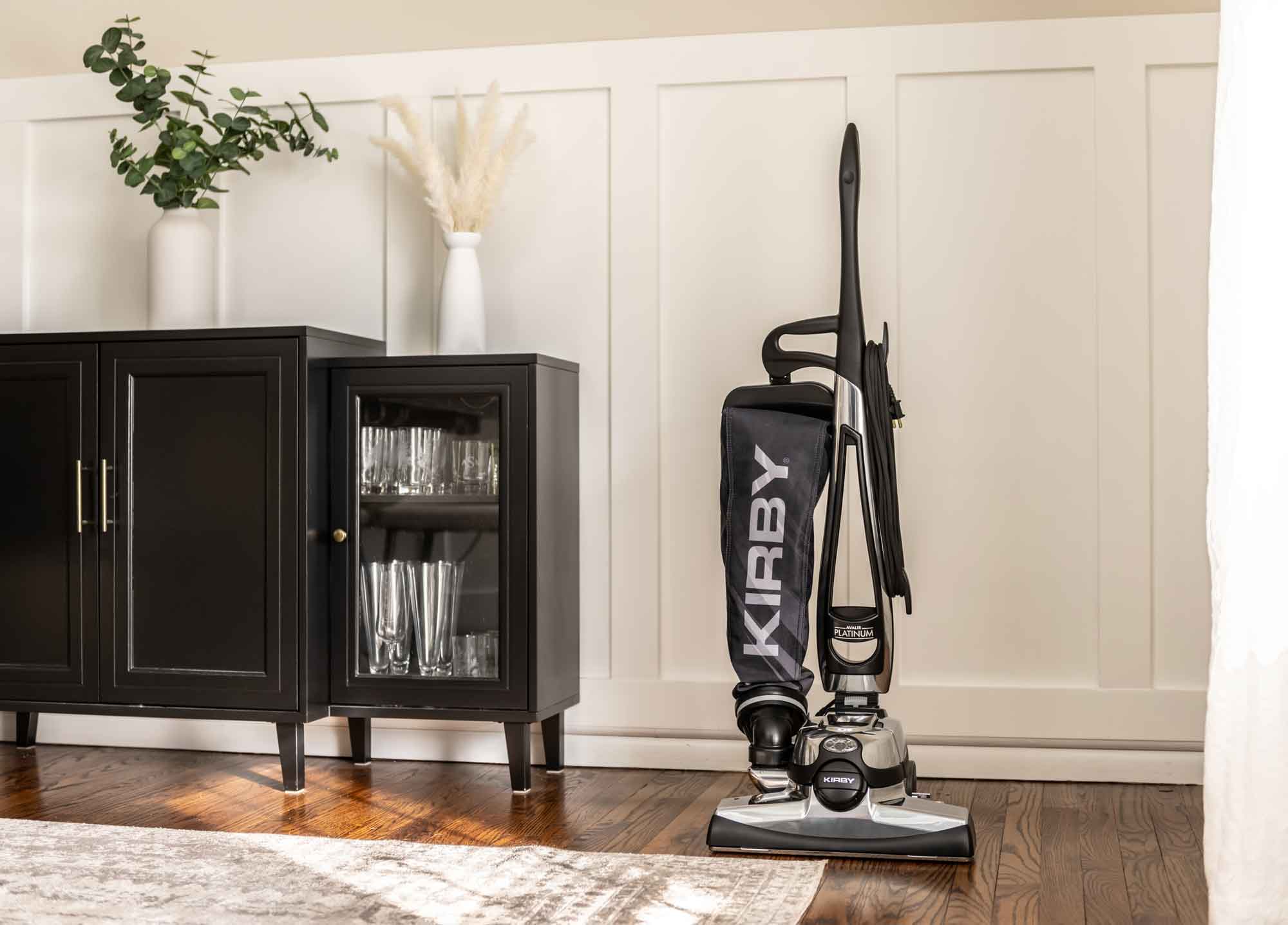 Kirby Avalir Platinum upright vacuum, black and silver, standing next to a black cabinet.