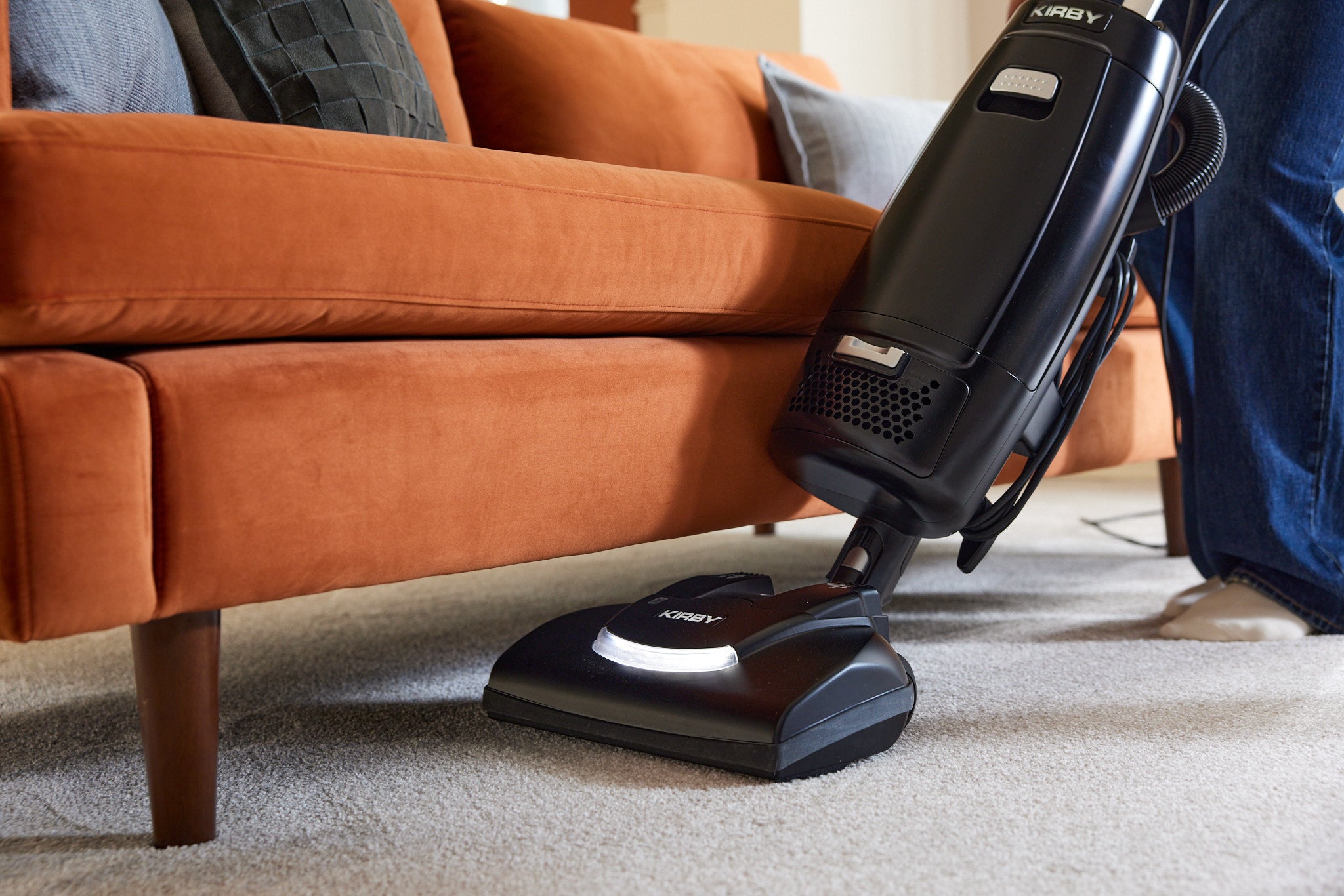 A black Kirby Signature Series V530 upright vacuum with a multi-surface Power Nozzle, shown being used under an orange couch on a light carpet.
