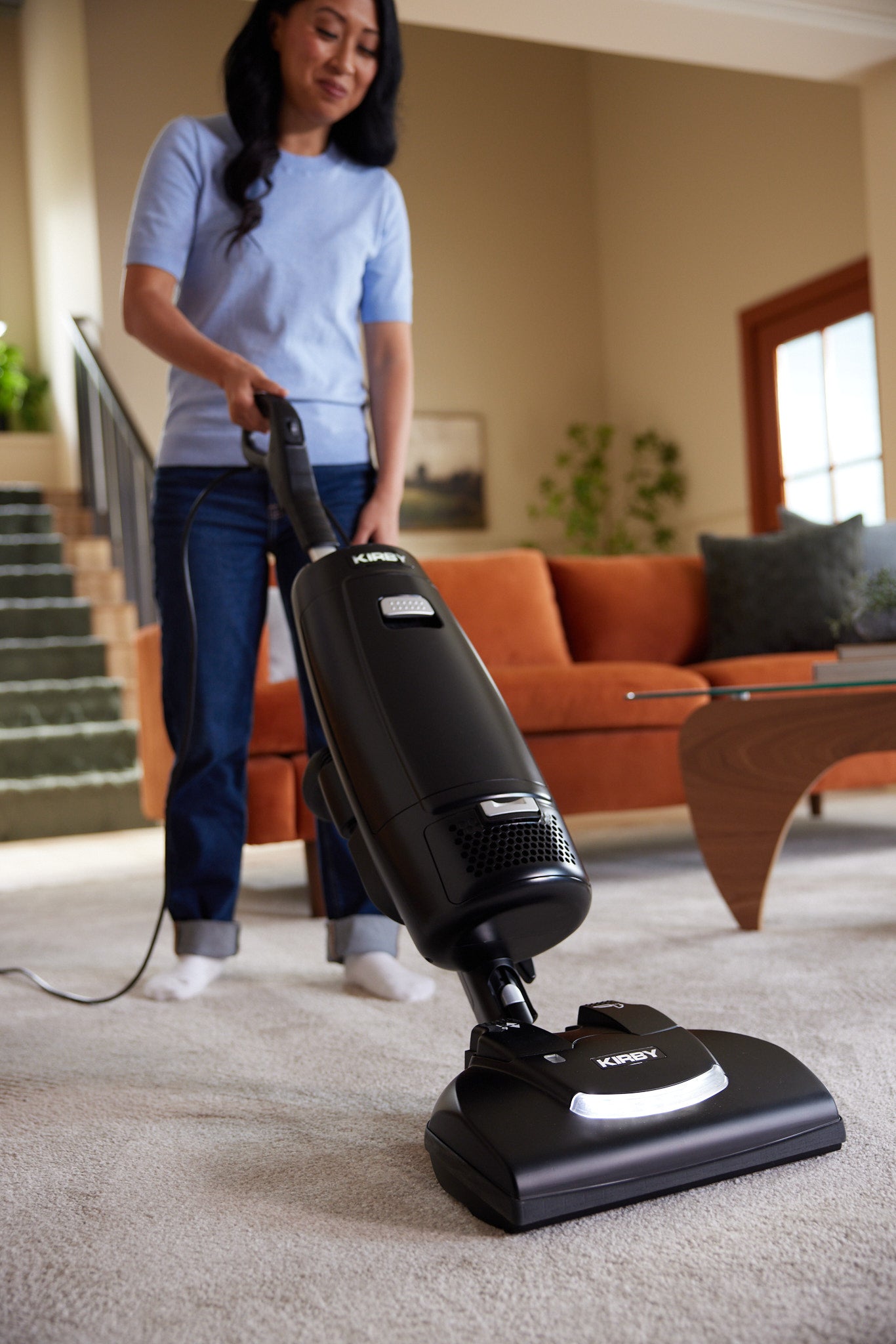 A woman in a living room using a black Kirby Signature Series V530 upright vacuum on a beige carpet, with an orange sofa and green staircase in the background.