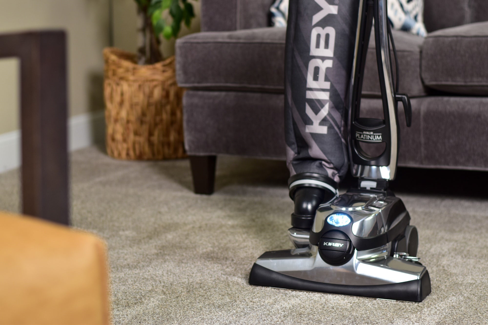 Kirby Avalir Platinum vacuum cleaner in black and silver, cleaning a carpet next to a gray sofa.