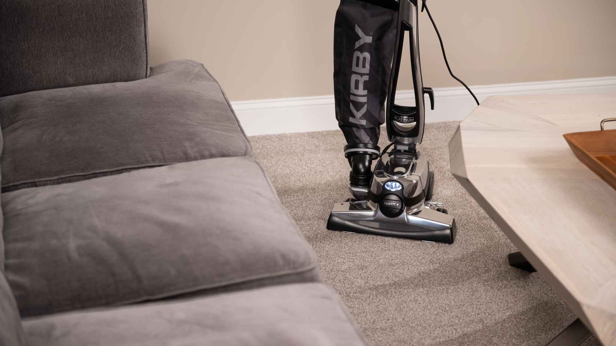 Kirby Avalir Platinum vacuum cleaner in black and silver, cleaning a carpet next to a gray sofa.