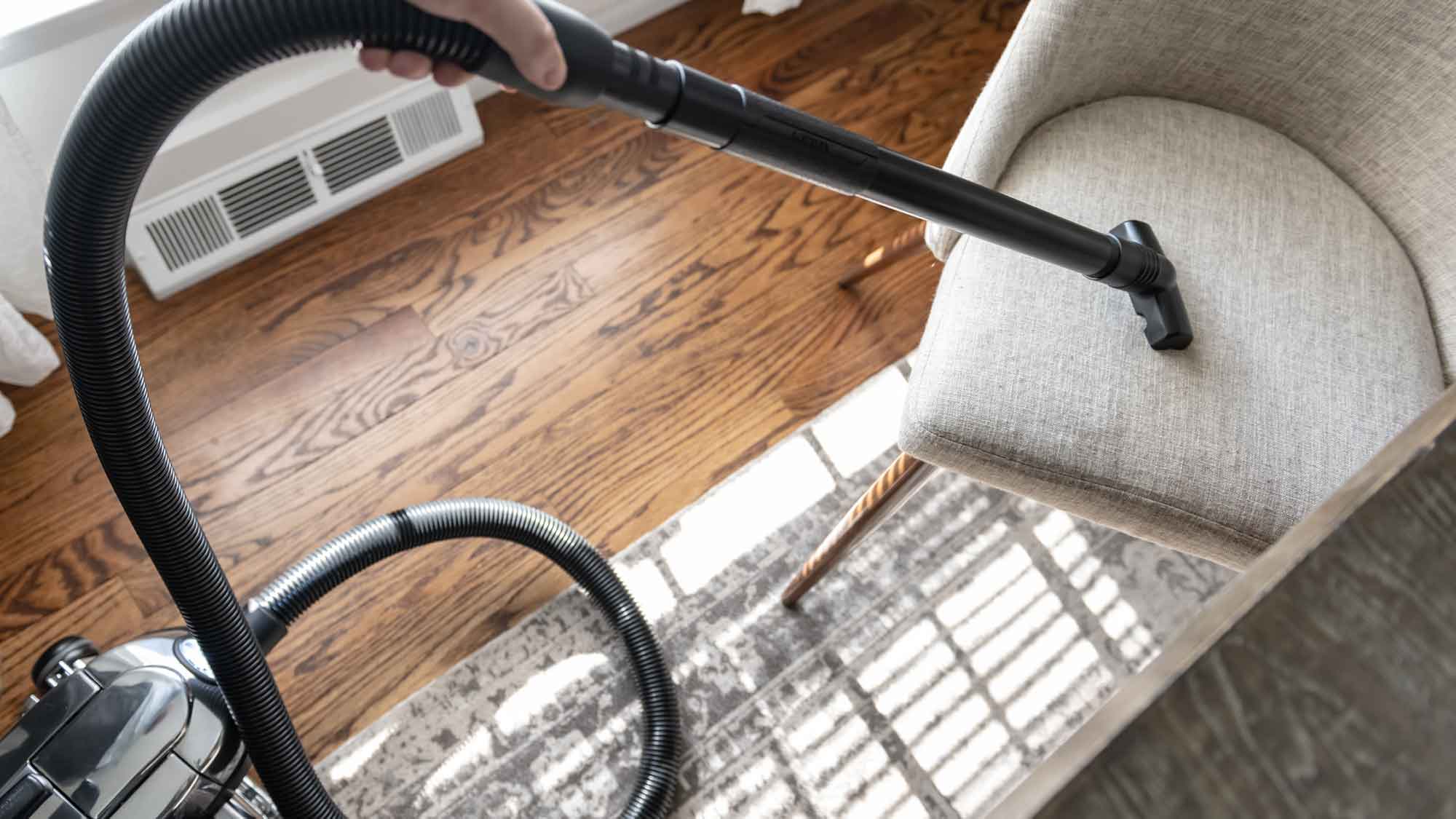 A person using a Kirby vacuum upholstery attachment to clean a chair in a living room