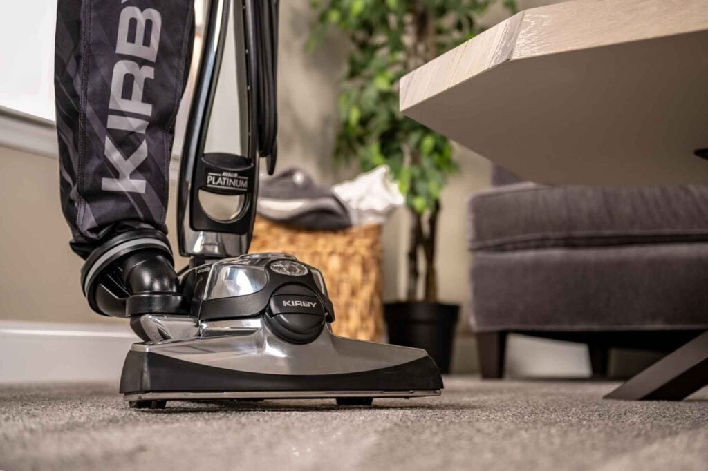 Kirby Avalir Platinum vacuum cleaner in silver and black, shown on a carpet near a coffee table.