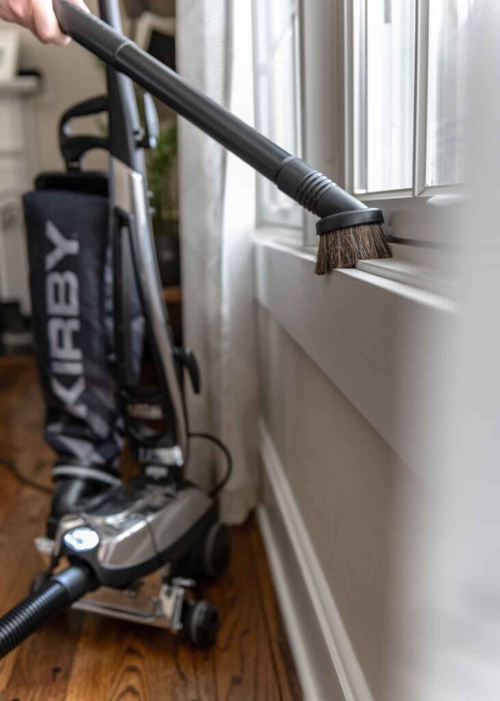 Kirby vacuum with a black and silver design, using a dusting brush attachment on a window sill.