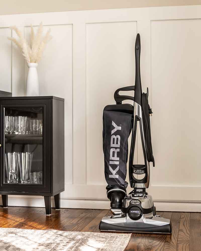 Kirby Avalir Platinum upright vacuum, black and silver, standing next to a black cabinet.