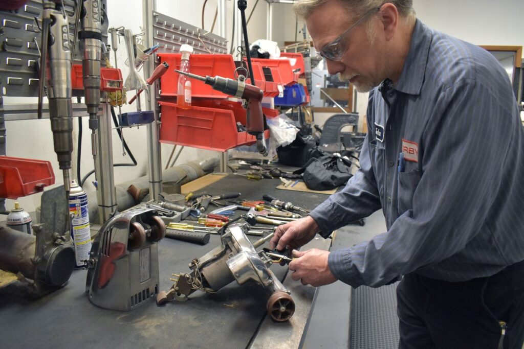 Technician repairing a silver Kirby vacuum on a workbench with various tools.