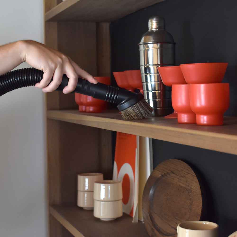 Hand using a Kirby vacuum attachment to clean a wooden shelf with red and beige cups and a silver shaker.