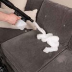 Person using a black Kirby portable sprayer to apply foam on a gray couch.